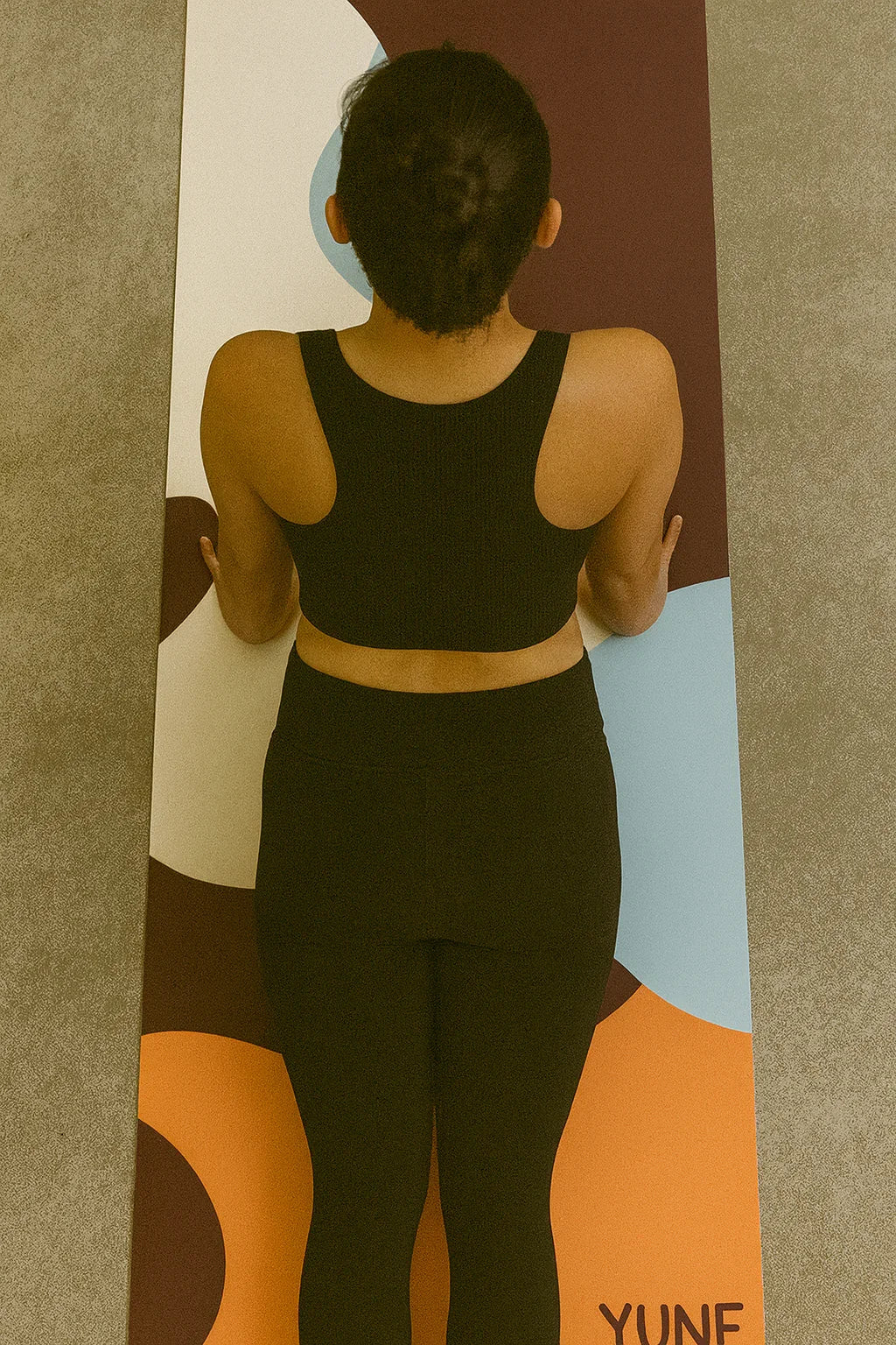 Person wearing a black athletic outfit standing on a colorful yoga mat with 'Yuna' branding.
